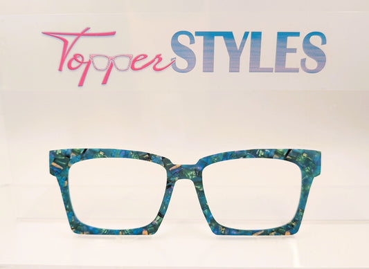 A pair of square-frame eyeglasses with a marbled teal, blue, and gold acetate frame, displayed on a white shelf against a white background with a 'Topper STYLES' logo sign above.