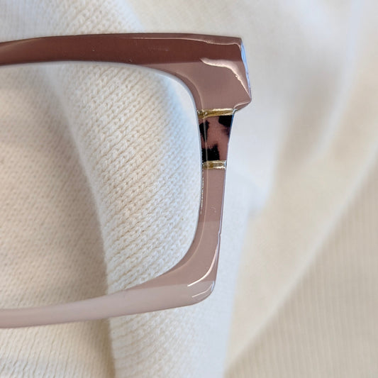 A close-up of a pair of eyeglasses with a glossy taupe-brown acetate frame resting on a textured cream knit fabric background.