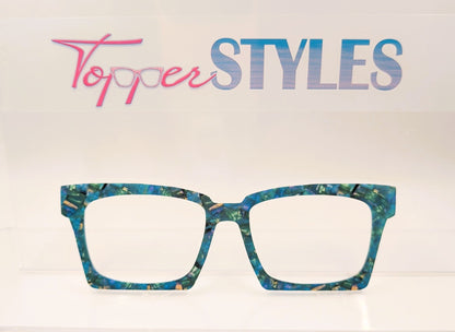 A pair of square-frame eyeglasses with a marbled teal, blue, and gold acetate frame, displayed on a white shelf against a white background with a 'Topper STYLES' logo sign above.